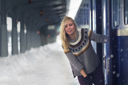 front view of blonde beautiful Caucasian young woman with long hair and warm sweater smiling near the door of a passenger train in a railway station, snowy, winter time の写真素材