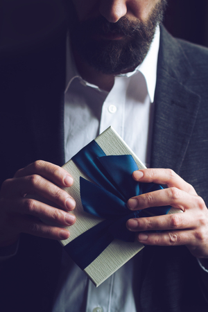 Horizontal close up of Caucasian man with beard in black suit and white shirt holding a gift box with blue large ribbon natural lightの写真素材