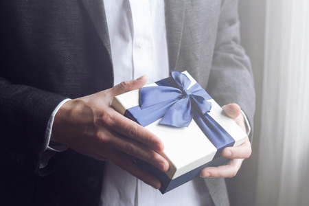 Horizontal close up of Caucasian man in black suit and white shirt offering a gift box with blue large ribbon selective focus low key dark natural lightの写真素材