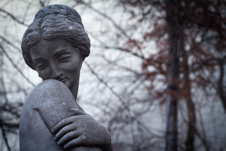 front view closeup detail of dark woman stone statue covering her breasts with one hand with bare trees branches in the backgroundの写真素材