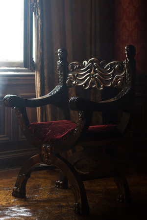 side view of antique armchair with round shape and wooden carved arms in the corner of a classical style room with vintage red wall paper natural lightningの写真素材