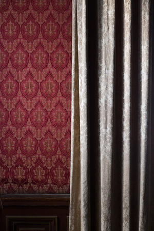 front view close up detail of light cream colored curtain covering red wallpaper wall with classic ornamentsの写真素材