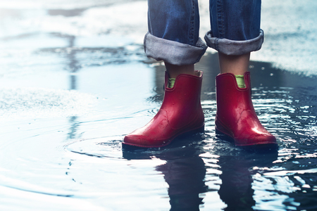 front view closeup of young Caucasian woman feet with red short rubber boots and blue jeans standing in a puddle of water after rain on a city streetの写真素材