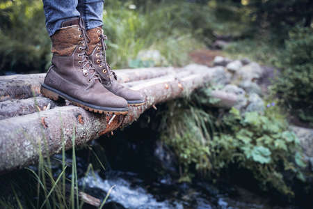 perspective view closeup of woman with brown tall leather hiking boots and grey jeans standing on a wooden trunk tree bridge over a mountain water stream in the forestの写真素材