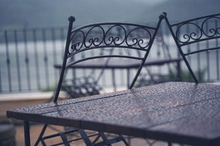 closeup detail of black iron metal chairs near wooden table with rain drops on rainy outdoor...