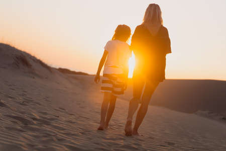 rear view of young woman and her child wearing shorts and towards the horizon on sand dunes at sunrise with sun shining between their silhouettesの写真素材