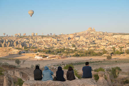 GOREME, TURKEY - AUGUST 5, 2021: People watch in the morning sitting on rocks as hot air balloons fly the tourists over the Cappadocia valley with Uchisar Castle in the backgroundのeditorial素材