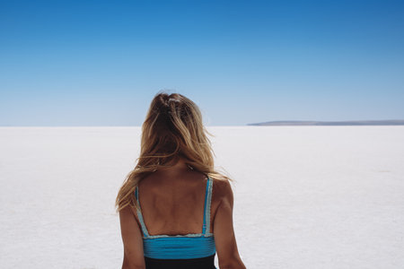 rear view of blonde beautiful Caucasian young woman dressed in blue looking at white salt lake stretching to the horizon with clear sky aboveの写真素材