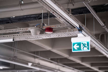 front view closeup of lighted green exit sign hanging on building pipes and pointing the way out of...