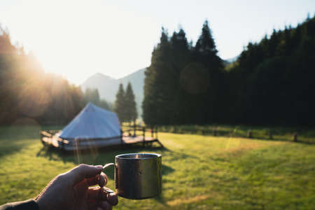 closeup front view hand holding a metallic coffee cup in the morning with glamping tent camping ground in the background in a green mountain meadow surrounded by fir tree forestの写真素材