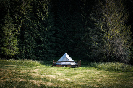 front view of secluded glamping camping tent with white cotton fabric on wooden deck in green meadow surrounded by fir tree forestの写真素材