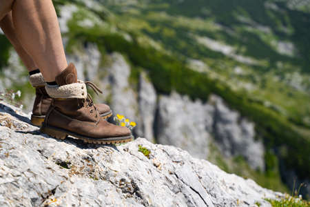 side view closeup detail of Caucasian woman legs with light colored shorts and hiking brown leather boots taking a break on mountain rockの写真素材