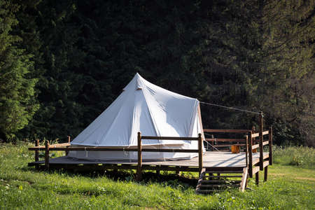 side view of secluded glamping camping tent with white cotton fabric on wooden deck in green meadow surrounded by fir tree forestの写真素材