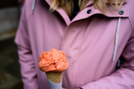 A vibrant orange gelato cone held by a person wearing a pink coat in an outdoor setting