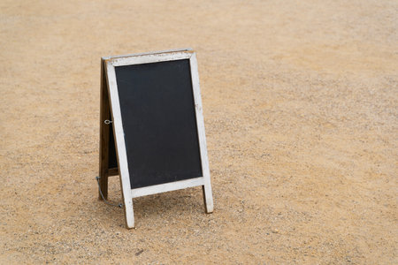 A rustic blackboard stand with a white wooden frame placed outdoors on a sandy ground surface for signage or messagingの写真素材