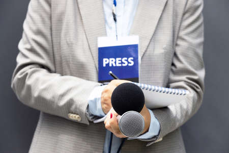 Female journalist at news conference or media event, writing notes, holding microphone. Journalism concept.の写真素材