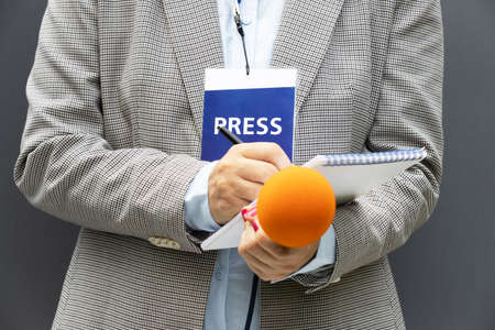 Female journalist at news conference or media event, writing notes, holding microphone. Journalism concept.の写真素材