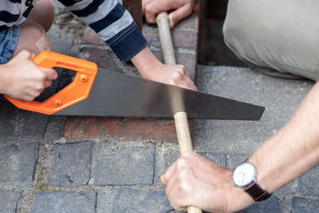 Boy is sawing a wooden stick that is being held by his fatherの写真素材