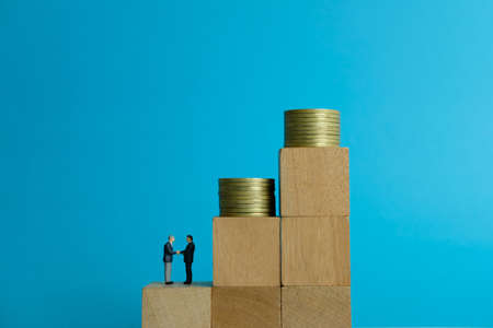 Miniature business concept - two businessman make an handshake for partnership above wooden puzzle and coin stackの写真素材