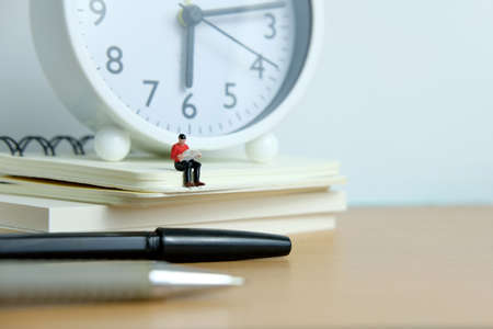 Young man sitting on book pile while reading with pen and white clock