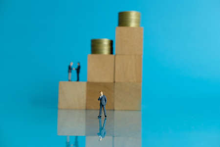 Miniature business concept - thinking businessman standing in front of wooden block with coin stack...