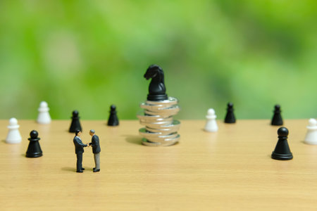 Business strategy conceptual photo - miniature businessman make handshake partnership agreement in front of coin stack with chess pawn on top of itの写真素材