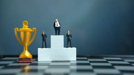 Miniature business concept - businessman standing on white winner podium with golden and silver trophyの写真素材