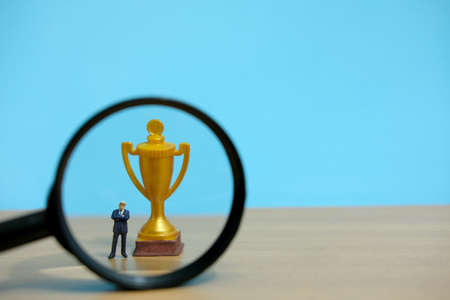 Miniature business concept - businessman standing in front of golden trophy and magnifier glassの写真素材