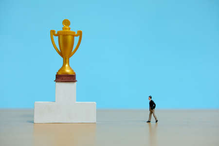 Miniature business concept - businessman walking toward golden trophy above white podiumの写真素材