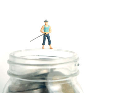 Miniature people toy photography. An angler standing above money jar fishing coin. Isolated on white...