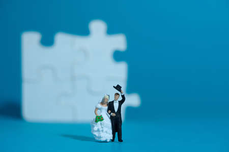 Bride and groom married miniature people stand in front of white puzzle jigsaw, isolated cut out on a blue background. Wedding plan strategy and choice. Image photoの写真素材