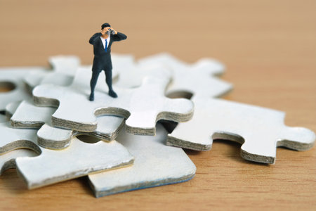 Businessmen standing above jigsaw puzzle piece stack using binoculars above wooden table. Miniature tiny people toys photography.の写真素材