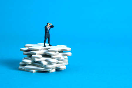Businessmen standing above jigsaw puzzle piece stack using binoculars above wooden table. Miniature tiny people toys photography. isolated on blue background.の写真素材