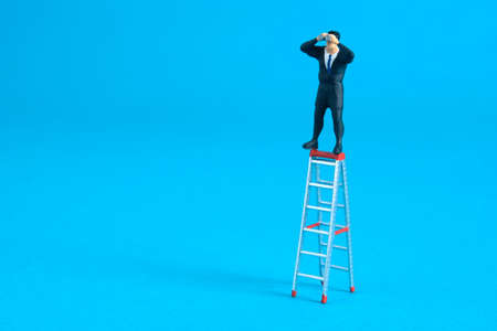 Businessman using binoculars standing above folding ladder. Miniature tiny people toys photography. isolated on blue background.の写真素材