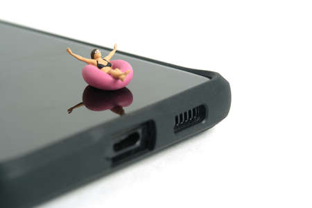 Miniature people toy figure photography. Virtual travel concept, girl swimming with rubber tube ring above smartphone, isolated on white background. Image photoの写真素材