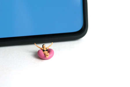 Miniature people toy figure photography. Virtual travel concept, girl swimming with rubber tube ring above smartphone, isolated on white background. Image photoの写真素材