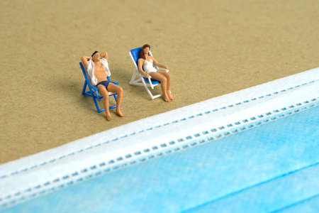 Miniature people toy figure photography. Safety travel concept, Men and girl relaxing at beach chair in front of blue face mask. Image photoの写真素材