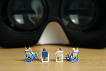 Miniature people toy figure photography. Virtual travel concept, group of men and girl relaxing seat while watching in front of goggles device, isolated on white background. Image photoの写真素材