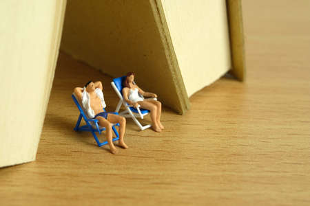 Miniature people toy figure photography. Men and girl couple relaxing seat at beach chair under book tent. Image photoの写真素材
