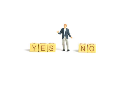 Miniature people toy figure photography. A shrugging businessman standing in the middle of yes and no  wooden cube block. Isolated on white background. Image photoの写真素材