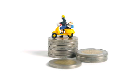 Miniature people toys conceptual photography. Cash on delivery and shipping cost service. Postman courier standing above money coin stack, isolated white background.の写真素材