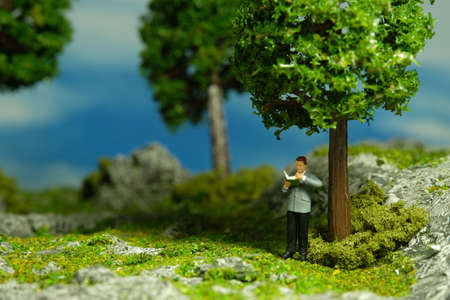Miniature people toy figure photography. A men standing under a tree while reading a book, relaxing at garden. Image photoの写真素材