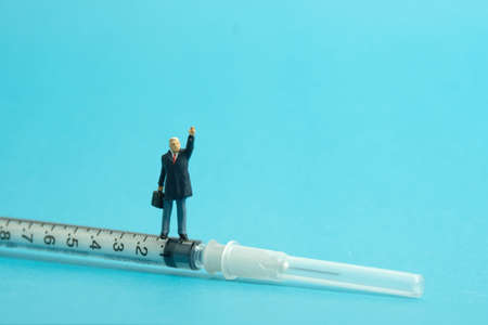 Miniature people toy figure photography. Vaccination encouragement concept. A businessman standing above syringe vaccine while raise his hand. Isolated on blue background. Image photoの写真素材