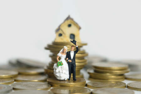 Housing for newlywed concept miniature people, toys photography. Bride and groom standing above coin pile in front of mini house isolated on white background. Image photoの写真素材