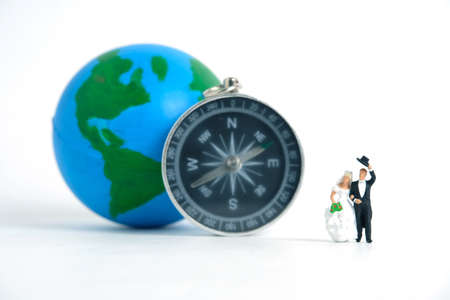 Honeymoon location selection concept miniature people, toys photography. Bride and groom standing in front of globe and compass isolated on white background. Image photoの写真素材