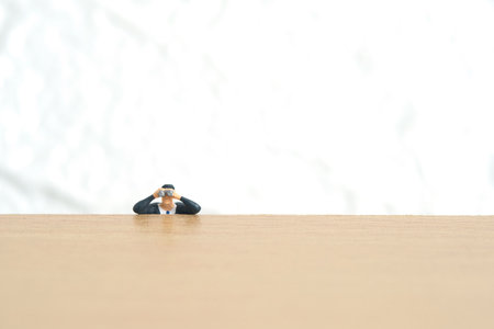 Businessman using binoculars hiding on wooden desk. Miniature tiny people toys photography. isolated on white background.の写真素材