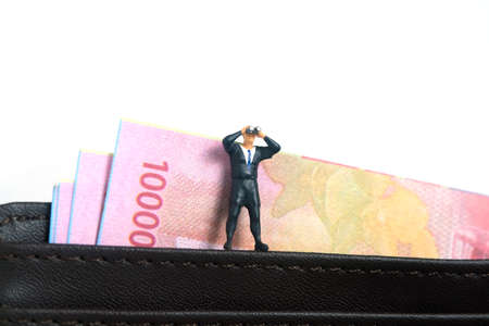 Businessman using binoculars standing on leather wallet with money inside. Miniature tiny people...