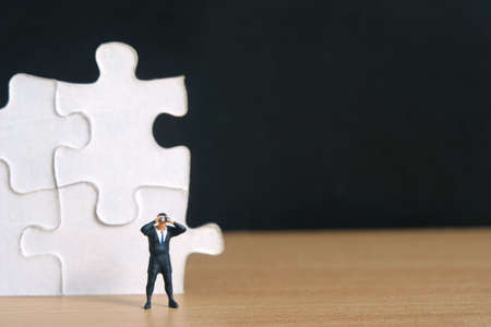Businessmen standing in front of jigsaw puzzle piece stack using binoculars on wooden table. Miniature tiny people toys photography.の写真素材