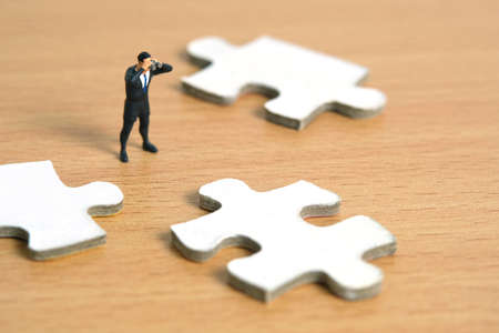 Businessmen standing in between three jigsaw puzzle piece using binoculars above wooden table. Miniature tiny people toys photography.の写真素材