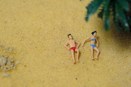 Miniature people toy figure photography. Top eagle view of men and girl couple relaxing, lying at beach sand when daylight at seaside. Image photoの写真素材